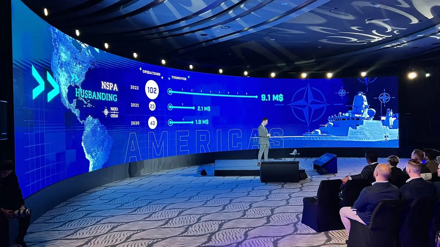 Relatore su palco davanti a uno schermo curvo con mappe e dati