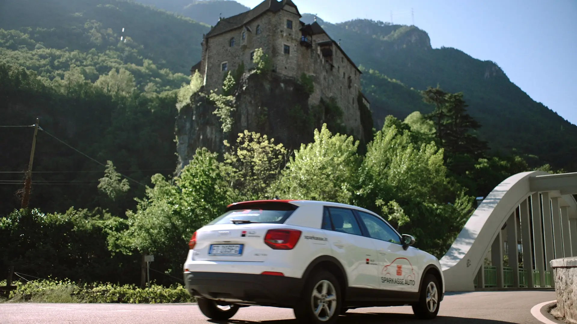 Auto con i colori Sparkasse su una strada di montagna, con un castello sulla rupe