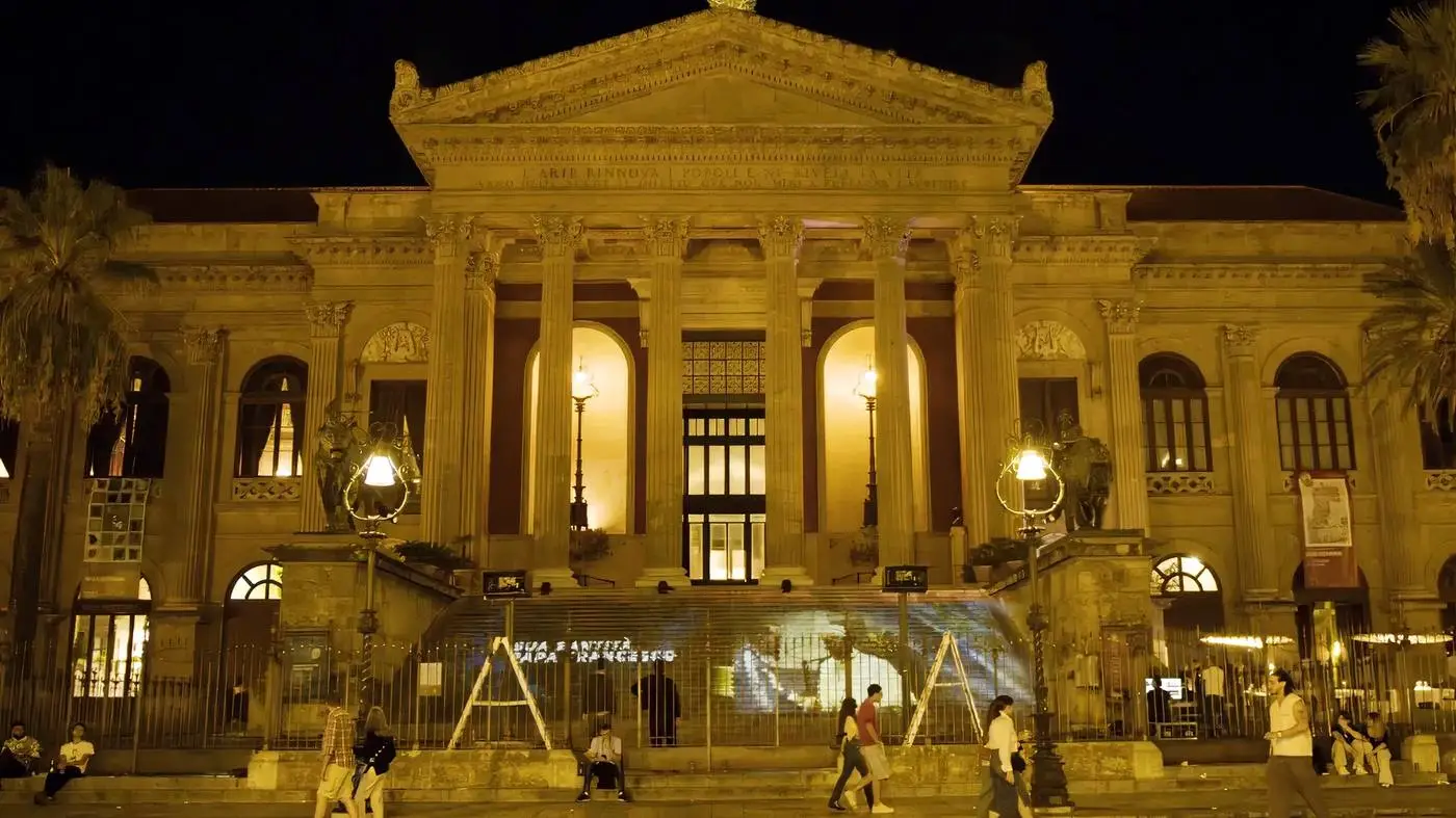 Facciata di un teatro neoclassico illuminata di notte con pubblico sulla scalinata